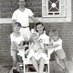 Father and 2 young sons stand; wife sits with baby on her lap-brick building in background