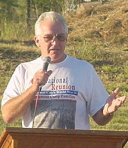 man speaking at podium wearing Crystal City, TX Internment Camp Reunion T-shirt
