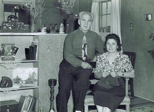 couple seated in corner; flowers, books, vases, etc on shelves to their right
