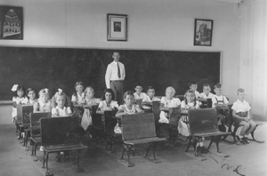 class room, with children at school desks and teacher at back of room