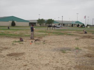 3 people, (1 standing, 2 on their knees) in large, dirt and grass area