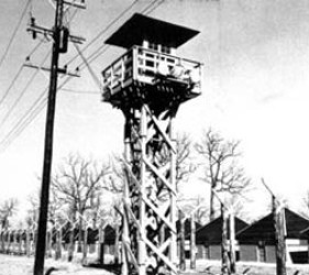 photograph of guard tower, with barbed wire and huts in background