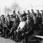 group of internees on small bleachers, with barbed wire fence behind them