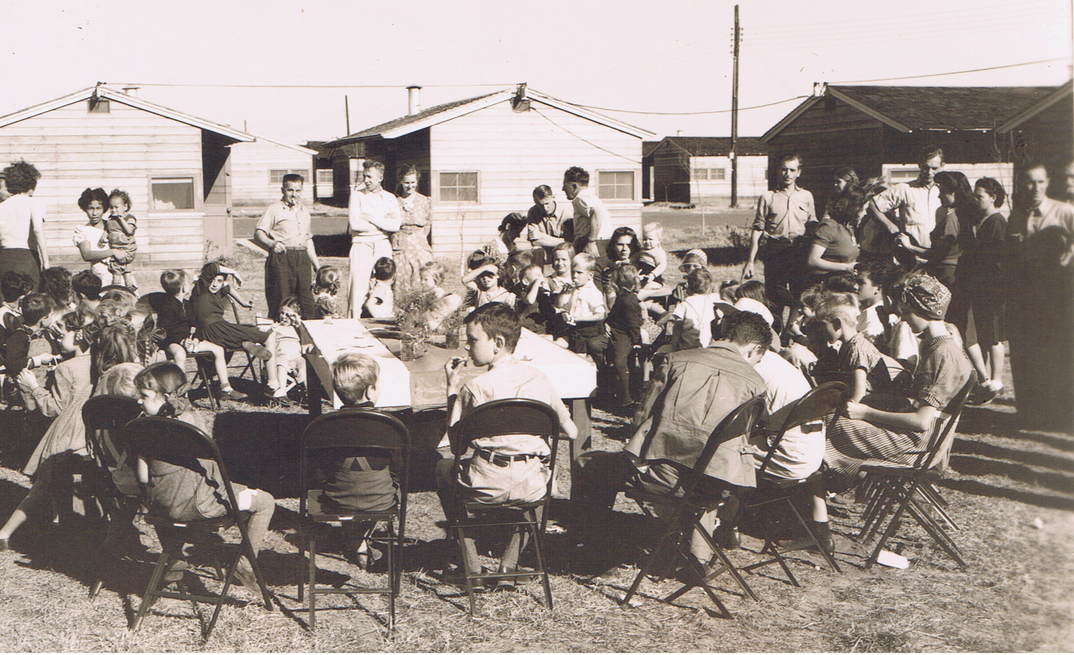 children and adults around outdoor table with living huts in background